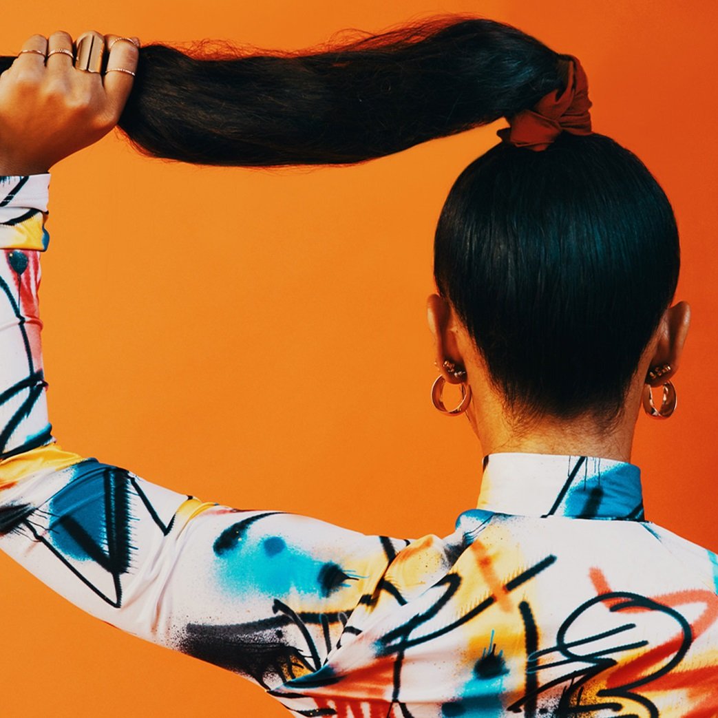 A person with long, dark hair holds their high ponytail against an orange background.